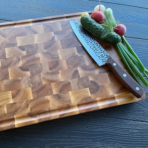 Peut inclure: Une planche à découper rectangulaire en bois de bout avec un motif de grain naturel brun et un bord rainuré. Un couteau de cuisine avec une lame en métal martelé et un manche en bois brun repose sur la planche à côté d'un concombre et de radis.