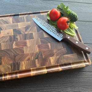 May include: A rectangular end-grain wooden cutting board with a dark brown, multi-toned block pattern and a grooved perimeter. A chef's knife with a hammered metal blade and a brown wooden handle rests on the board next to two red tomatoes.