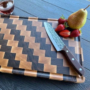 May include: A rectangular wooden cutting board with a dark and light brown checkered pattern, featuring a grooved edge. A silver hammered-finish knife with a dark wood handle rests on the board next to a green pear and three red strawberries.