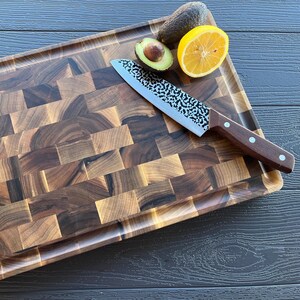 May include: A rectangular end-grain walnut wood cutting board with a dark brown checkered pattern and a grooved perimeter. A chef's knife with a hammered metal blade and a dark wood handle rests on the board next to a halved avocado and a lemon slice.