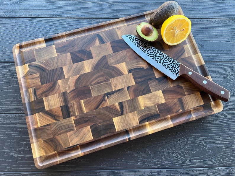 May include: A rectangular end-grain walnut wood cutting board with a dark brown checkered pattern and a grooved perimeter. A chef's knife with a hammered metal blade and a dark wood handle rests on the board next to a halved avocado and a lemon slice.