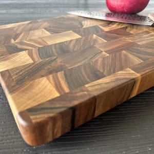 May include: A rectangular end-grain cutting board constructed from blocks of dark and light brown wood with a visible grain pattern. A stainless steel kitchen knife with a brown handle and a red apple are positioned on the surface of the board.