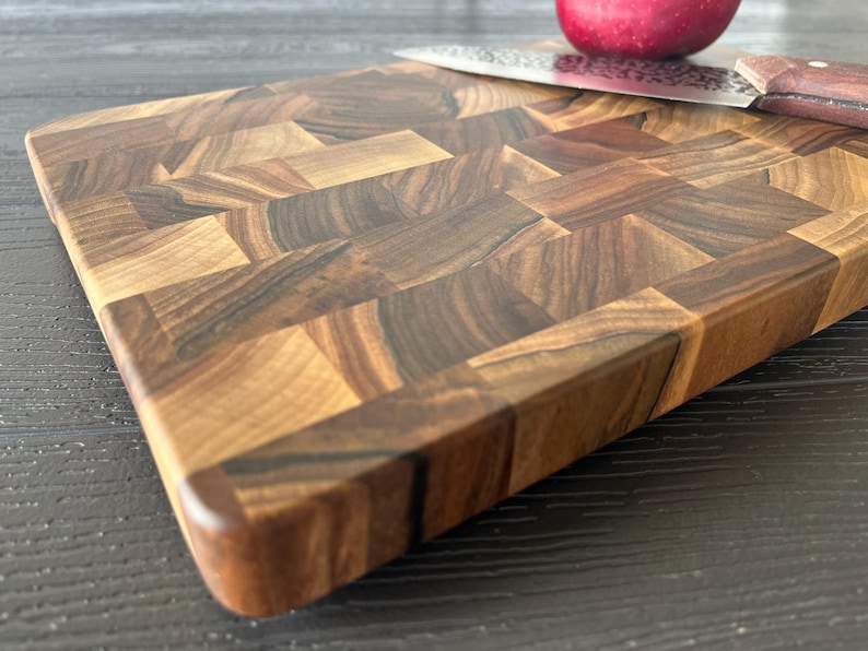 May include: A rectangular end-grain cutting board constructed from blocks of dark and light brown wood with a visible grain pattern. A stainless steel kitchen knife with a brown handle and a red apple are positioned on the surface of the board.