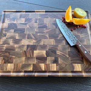 K&ouml;nnte beinhalten: Ein rechteckiges Stirnholz-Schneidebrett mit dunkelbraunem Schachbrettmuster und einer Saftrille am Rand. Ein Kochmesser mit geh&auml;mmertem Metallblatt und braunem Holzgriff liegt auf dem Brett neben Orangenscheiben und einer Kiwischeibe.