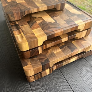 K&ouml;nnte beinhalten: Ein Stapel aus drei rechteckigen Schneidebrettern aus Holz mit Hirnholz-Konstruktion und einem Mosaikmuster aus hellen und dunkelbraunen Holzbl&ouml;cken. Jedes Brett hat abgerundete Ecken und eine umlaufende Saftrille am oberen Rand.