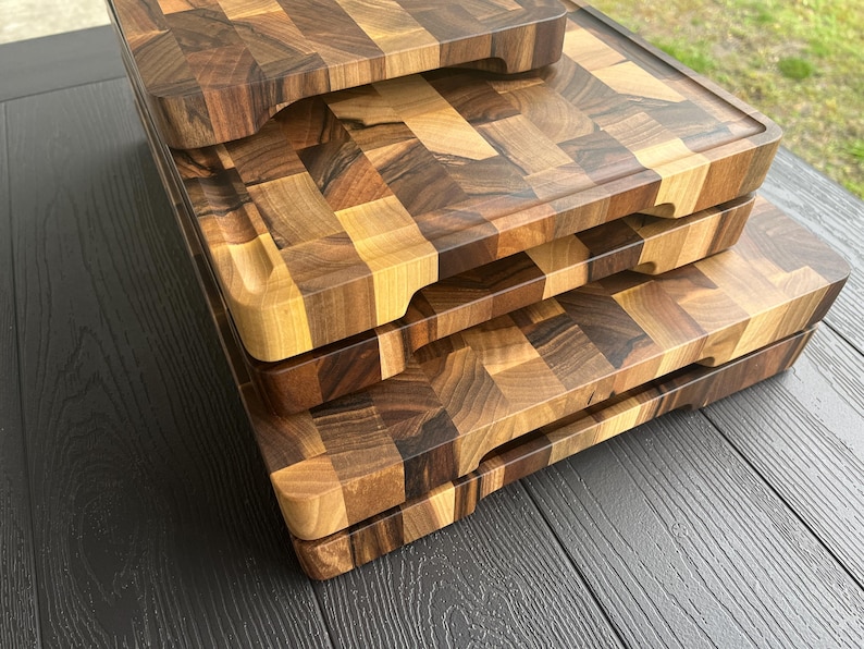 K&ouml;nnte beinhalten: Ein Stapel aus drei rechteckigen Schneidebrettern aus Holz mit Hirnholz-Konstruktion und einem Mosaikmuster aus hellen und dunkelbraunen Holzbl&ouml;cken. Jedes Brett hat abgerundete Ecken und eine umlaufende Saftrille am oberen Rand.