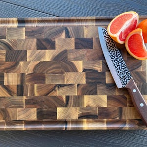 May include: A rectangular end-grain wooden cutting board with a dark brown checkered pattern. A kitchen knife with a hammered metal blade and a brown wooden handle rests on the board next to one whole orange and two slices of pink grapefruit.