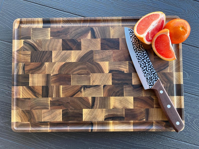 May include: A rectangular end-grain wooden cutting board with a dark brown checkered pattern. A kitchen knife with a hammered metal blade and a brown wooden handle rests on the board next to one whole orange and two slices of pink grapefruit.