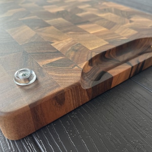 May include: A rectangular end-grain cutting board crafted from dark brown walnut wood with a visible checkerboard pattern. The board features a recessed handle carved into the side and two clear, circular rubber feet attached to the bottom surface.