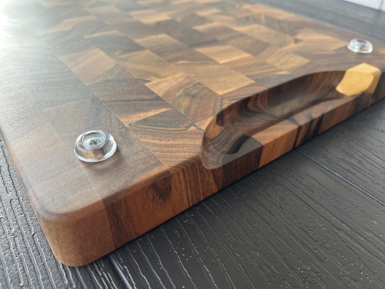 May include: A rectangular end-grain cutting board crafted from dark brown walnut wood with a visible checkerboard pattern. The board features a recessed handle carved into the side and two clear, circular rubber feet attached to the bottom surface.