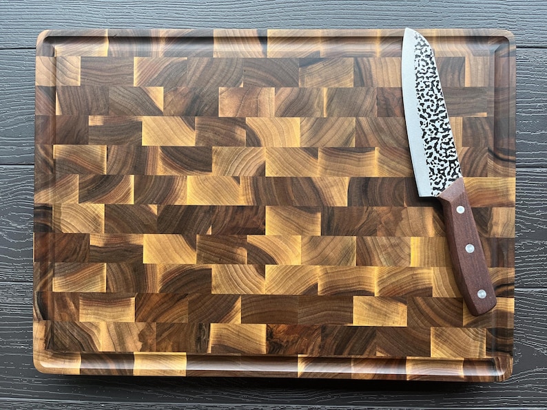 K&ouml;nnte beinhalten: Ein rechteckiges Stirnholz-Schneidebrett mit einem dunkel- und hellbraunen Schachbrettmuster und einer umlaufenden Saftrille. Ein K&uuml;chenmesser mit einer geh&auml;mmerten Metallklinge und einem braunen Holzgriff liegt auf der rechten Seite.