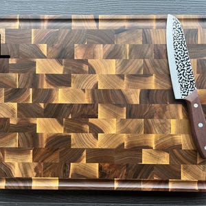 May include: A rectangular end-grain wooden cutting board with a dark and light brown checkered pattern and a juice groove along the perimeter. A kitchen knife with a hammered metal blade and a brown wooden handle rests on the board.