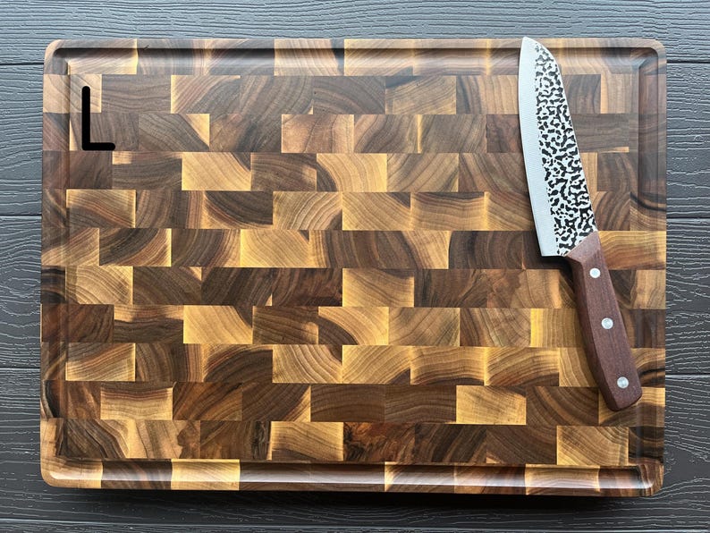 May include: A rectangular end-grain wooden cutting board with a dark and light brown checkered pattern and a juice groove along the perimeter. A kitchen knife with a hammered metal blade and a brown wooden handle rests on the board.