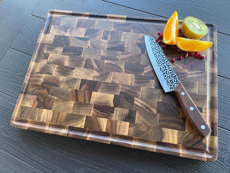 K&ouml;nnte beinhalten: Ein rechteckiges Stirnholz-Schneidebrett mit dunkelbraunem Schachbrettmuster und einer Saftrille. Ein Metall-Kochmesser mit geh&auml;mmertem Finish und braunem Holzgriff liegt zusammen mit Orangen- und Kiwischeiben auf dem Schneidebrett.