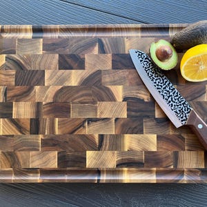May include: A rectangular end-grain wooden cutting board with a dark brown mosaic pattern. A chef's knife with a hammered metal blade and a brown wooden handle rests on the board, alongside a halved avocado and a slice of yellow lemon.