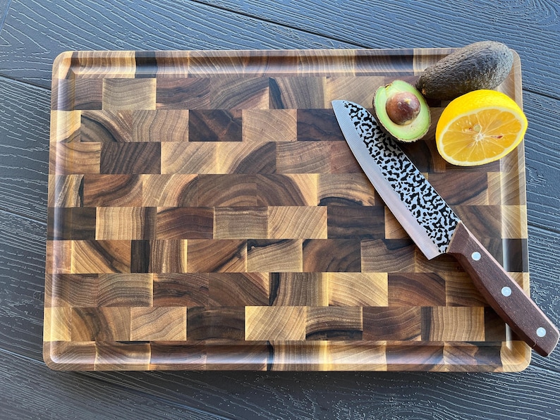 May include: A rectangular end-grain wooden cutting board with a dark brown mosaic pattern. A chef's knife with a hammered metal blade and a brown wooden handle rests on the board, alongside a halved avocado and a slice of yellow lemon.