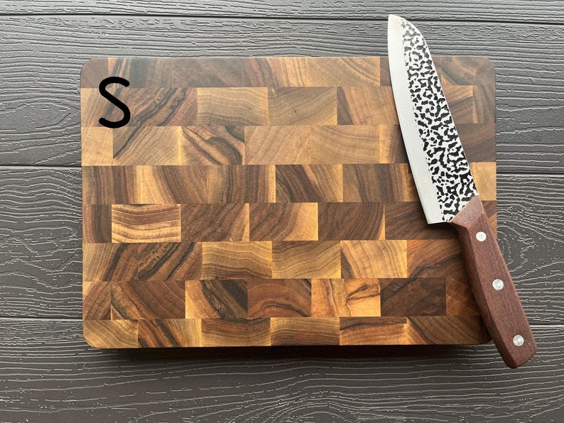 May include: A rectangular end-grain wooden cutting board with a dark brown, multi-toned block pattern and the letter S printed in black on the top left corner. A kitchen knife with a hammered metal blade and a brown wooden handle rests on top.