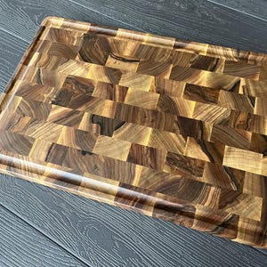 May include: A rectangular end-grain cutting board crafted from dark and light brown walnut wood blocks. The board features a smooth, polished surface with a distinct mosaic pattern of wood grain variations and a recessed juice groove perimeter.