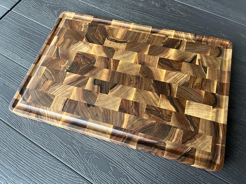 May include: A rectangular end-grain cutting board crafted from dark and light brown walnut wood blocks. The board features a smooth, polished surface with a distinct mosaic pattern of wood grain variations and a recessed juice groove perimeter.