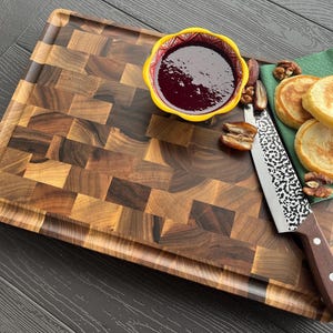 May include: A rectangular wooden cutting board with a dark brown end-grain checkerboard pattern. It features a yellow bowl filled with dark purple jam, a silver knife with a textured blade and brown handle, and two pancakes on a green napkin.