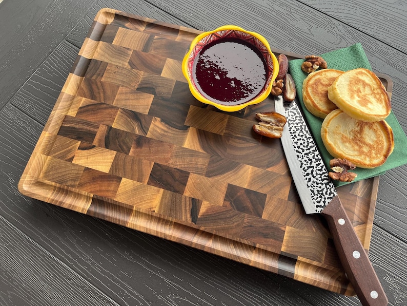 May include: A rectangular wooden cutting board with a dark brown end-grain checkerboard pattern. It features a yellow bowl filled with dark purple jam, a silver knife with a textured blade and brown handle, and two pancakes on a green napkin.