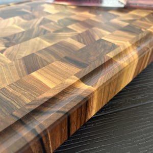 May include: A rectangular wooden cutting board featuring an end-grain construction with a mosaic of light and dark brown wood blocks. The board has a smooth, polished finish and a recessed juice groove running along the perimeter edge.