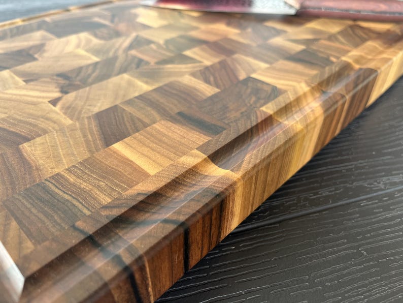 May include: A rectangular wooden cutting board featuring an end-grain construction with a mosaic of light and dark brown wood blocks. The board has a smooth, polished finish and a recessed juice groove running along the perimeter edge.
