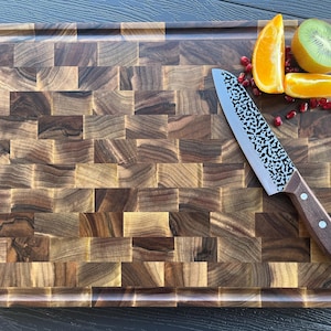 K&ouml;nnte beinhalten: Ein rechteckiges Stirnholz-Schneidebrett mit dunkelbraunem Schachbrettmuster. Ein K&uuml;chenmesser mit geh&auml;mmertem Metallblatt und braunem Holzgriff liegt auf dem Brett, zusammen mit Orangenscheiben, einer Kiwi und einigen Granatapfelkernen.