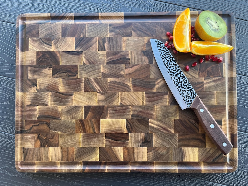 K&ouml;nnte beinhalten: Ein rechteckiges Stirnholz-Schneidebrett mit dunkelbraunem Schachbrettmuster. Ein K&uuml;chenmesser mit geh&auml;mmertem Metallblatt und braunem Holzgriff liegt auf dem Brett, zusammen mit Orangenscheiben, einer Kiwi und einigen Granatapfelkernen.
