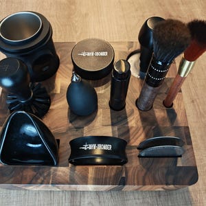 Puede incluir: Colección de accesorios de café negros, incluyendo un tamper, cepillos y un portafiltro, dispuestos sobre un bloque de madera. Los accesorios tienen un diseño elegante y moderno, con el texto "MWV-1BOMBER" visible.