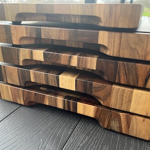 K&ouml;nnte beinhalten: Ein Stapel aus vier rechteckigen Holzschneidebrettern mit einer mehrfarbigen Maserung in hellen und dunklen Braunt&ouml;nen. Jedes Brett verf&uuml;gt &uuml;ber eine gefr&auml;ste horizontale Griffmulde an der Vorderkante und eine massive Holzkonstruktion.