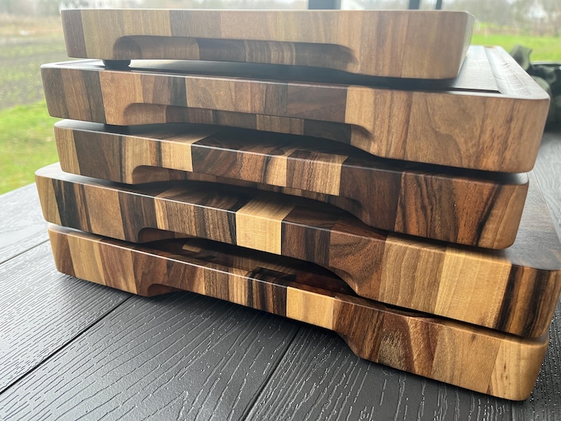 K&ouml;nnte beinhalten: Ein Stapel aus vier rechteckigen Holzschneidebrettern mit einer mehrfarbigen Maserung in hellen und dunklen Braunt&ouml;nen. Jedes Brett verf&uuml;gt &uuml;ber eine gefr&auml;ste horizontale Griffmulde an der Vorderkante und eine massive Holzkonstruktion.