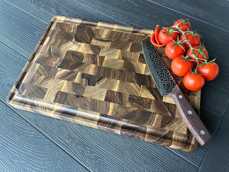 May include: A rectangular end-grain wooden cutting board with a dark brown, multi-tonal block pattern and a perimeter juice groove. A kitchen knife with a dark wood handle and a textured metal blade rests beside a cluster of fresh red tomatoes.