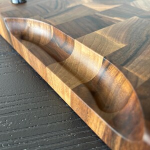 May include: A rectangular wooden cutting board featuring a dark and light brown checkered end-grain pattern. The board has a smooth, polished finish and includes a carved, recessed handle grip along the side for easy lifting and handling.