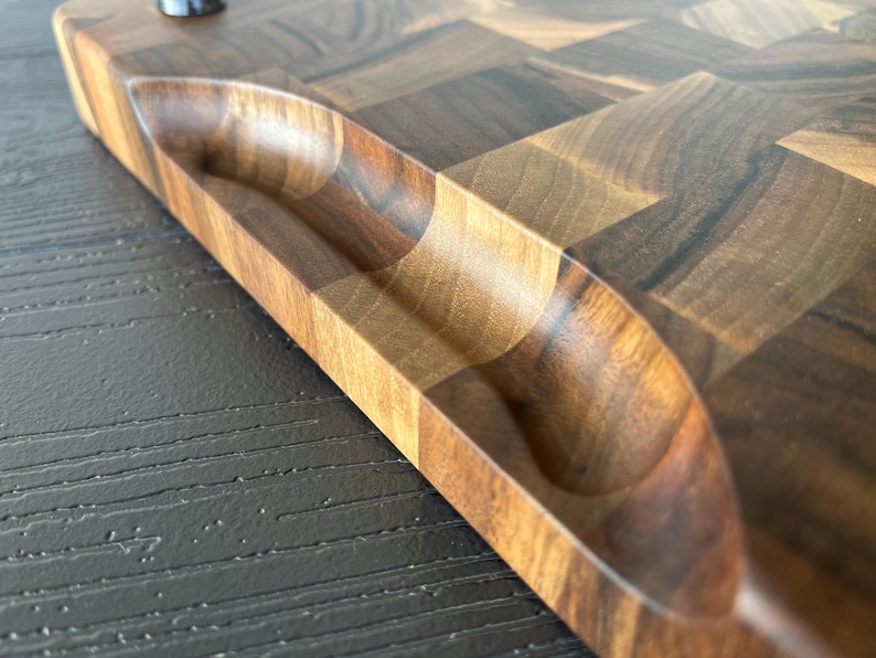 May include: A rectangular wooden cutting board featuring a dark and light brown checkered end-grain pattern. The board has a smooth, polished finish and includes a carved, recessed handle grip along the side for easy lifting and handling.