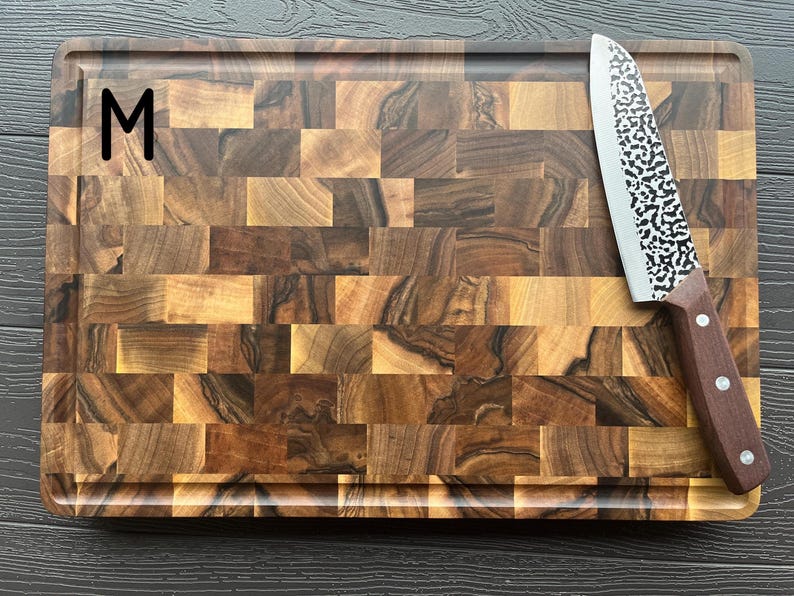 May include: A rectangular end-grain wooden cutting board with a dark and light brown checkered pattern and a juice groove along the perimeter. The letter M is printed in black on the top left. A chef knife with a hammered metal blade rests on top.