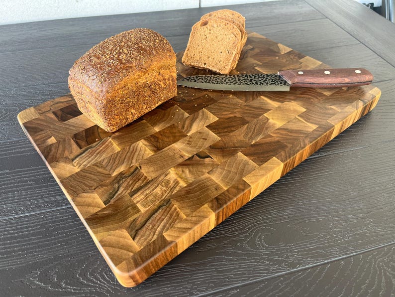 May include: A rectangular end-grain wooden cutting board with a dark brown checkered pattern. A loaf of brown bread sits on the board next to a few slices and a kitchen knife with a dark wooden handle and a patterned metal blade.