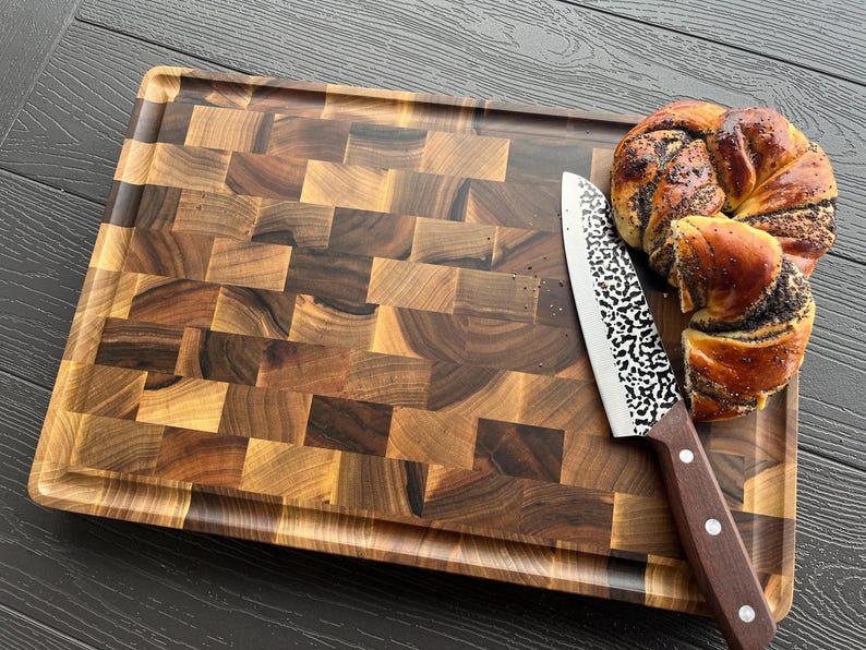 May include: A rectangular wooden cutting board with a dark and light brown checkered end-grain pattern, featuring a grooved perimeter. A kitchen knife with a hammered metal blade and a brown wooden handle rests on the board next to a poppy seed roll.