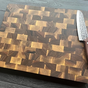 K&ouml;nnte beinhalten: Ein rechteckiges Stirnholz-Schneidebrett aus abwechselnd hell- und dunkelbraunen Holzbl&ouml;cken. Ein K&uuml;chenmesser mit einer geh&auml;mmerten Metallklinge und einem dunkelbraunen Holzgriff mit drei silbernen Nieten liegt auf der Oberfl&auml;che des Brettes.