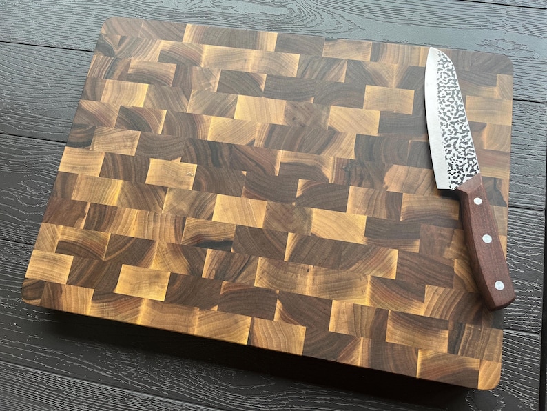 K&ouml;nnte beinhalten: Ein rechteckiges Stirnholz-Schneidebrett aus abwechselnd hell- und dunkelbraunen Holzbl&ouml;cken. Ein K&uuml;chenmesser mit einer geh&auml;mmerten Metallklinge und einem dunkelbraunen Holzgriff mit drei silbernen Nieten liegt auf der Oberfl&auml;che des Brettes.
