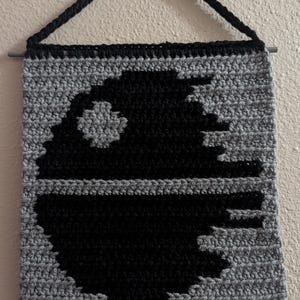 May include: A square crochet wall hanging featuring a black and gray spherical space station design. The piece is suspended from a horizontal rod with a black yarn hanger. The textured stitches create a detailed graphic of a large circular structure.