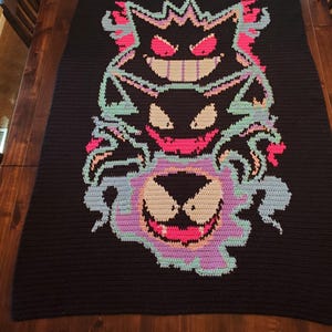 May include: A rectangular black crochet blanket featuring a vertical graphic design of three stacked ghost-like creatures. The figures are rendered in shades of neon pink, light blue, and cream, with sharp teeth and glowing eyes set against the dark background.