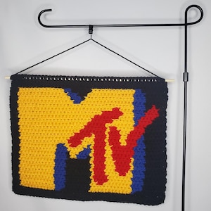 May include: A handmade, crocheted wall hanging featuring the iconic MTV logo. The design showcases a yellow background with a black border, and the MTV logo in red, blue, and yellow. The banner is suspended from a wooden dowel.