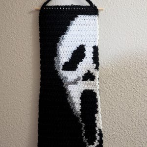 May include: A black and white crochet wall hanging depicting the Ghostface mask from the Scream film series. The mask is white against a black backdrop. The rectangular wall hanging is suspended from a wooden dowel.