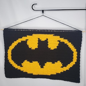 May include: A black and yellow crochet Batman logo wall hanging. The Batman symbol is yellow and centered on a black background. The wall hanging is rectangular and is hung from a black hanger.