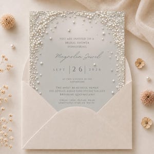Puede incluir: Una tarjeta de invitaci&oacute;n gris claro para una despedida de soltera en un sobre a juego, con un borde de perlas blancas y cristales transparentes. El texto dice: You are invited to a bridal shower honouring Magnolia Jewel, Sept 26 2026.