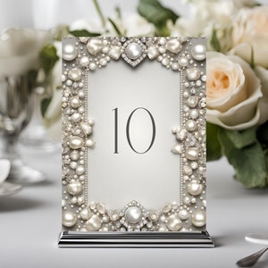 Puede incluir: Una tarjeta de número de mesa blanca con un borde de perlas y diamantes de imitación. El número 10 está impreso en negro en la tarjeta. La tarjeta está en un soporte plateado.