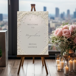 Puede incluir: Un caballete de madera exhibe un letrero con el texto "Welcome to Magnolia's Bridal Shower". El letrero tiene un borde y detalles de perlas. Rosas rosas y velas se ven en el fondo.