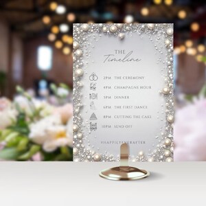 May include: A white wedding timeline sign with a pearl border. The sign lists the wedding day events with icons and times: 2 PM The Ceremony, 4 PM Champagne Hour, 5 PM Dinner, 6 PM The First Dance, 8 PM Cutting the Cake, 10 PM Send Off. The sign also includes the hashtag #HappilyEverAfter.