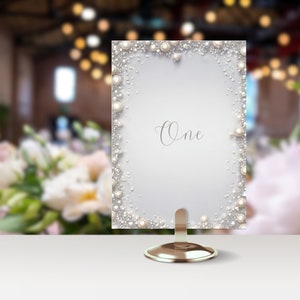 May include: A white table number card with a pearl border and the word "One" written in cursive. The card is on a gold metal stand.